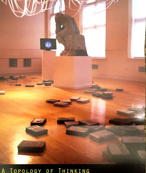 Exhibit card showing installation with objects on floor, a video screen and a sculpture like Rodin's thinker