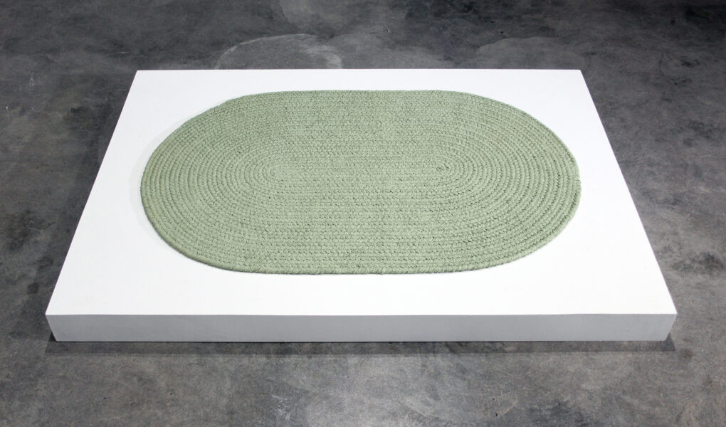 A round form reminiscent of a green coiled rug on a white plinth low to cement floor