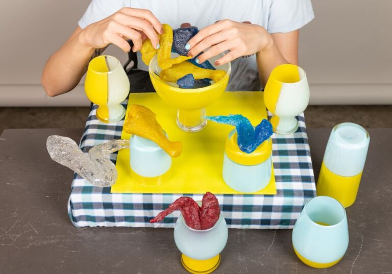 Photograph of place setting with yellow and light blue cups and bowl with colored glass chicken legs and a person's hands visible
