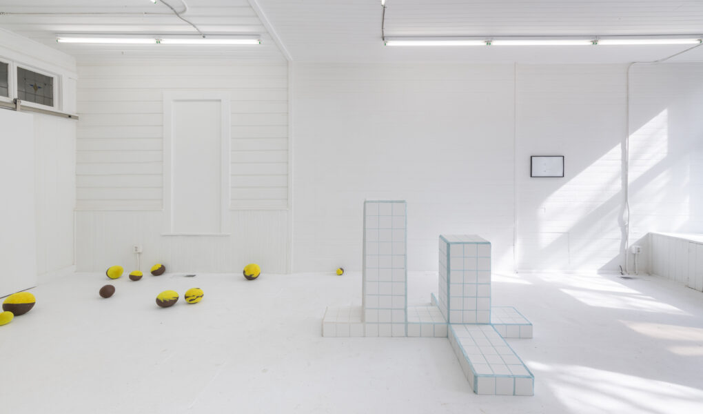 photo of artwork installation in very white room including white pedestals and small round yellow objects on floor